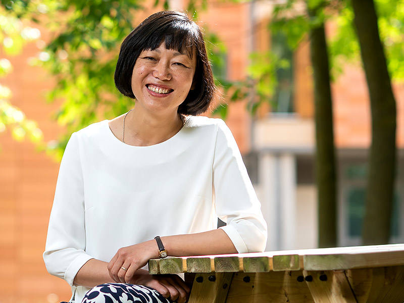 Bo Hu sits smiling surrounded by trees and other greenery outside the University of Oxford China Centre based at St Hugh’s College