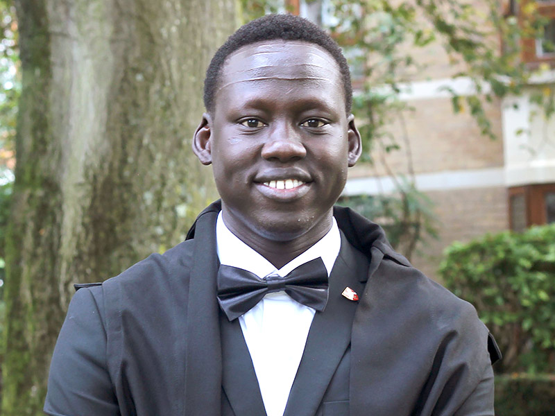 Lam Joar stands outside a college building wearing academic dress