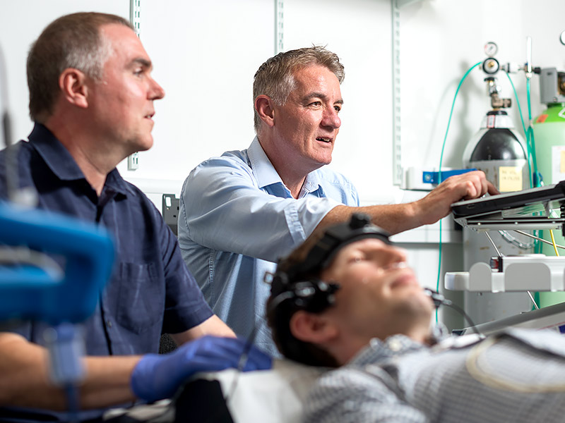 Clinical setting where a researcher takes observations of a patient and Professor Peter Rothwell looks on