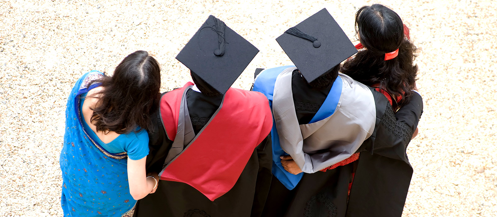 Two graduating students wearing academic dress stand outside with family members