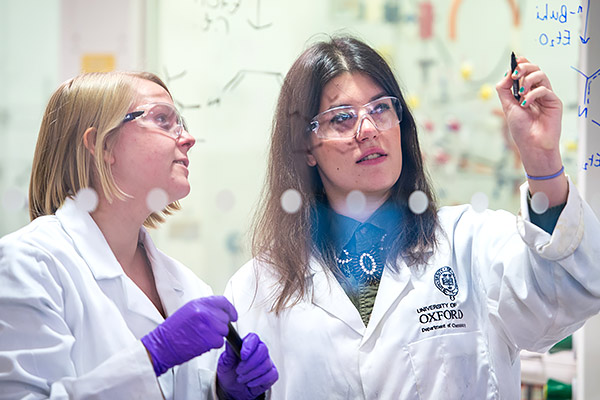 Two students wearing lab coats write out formulas on a windowpane in the chemistry