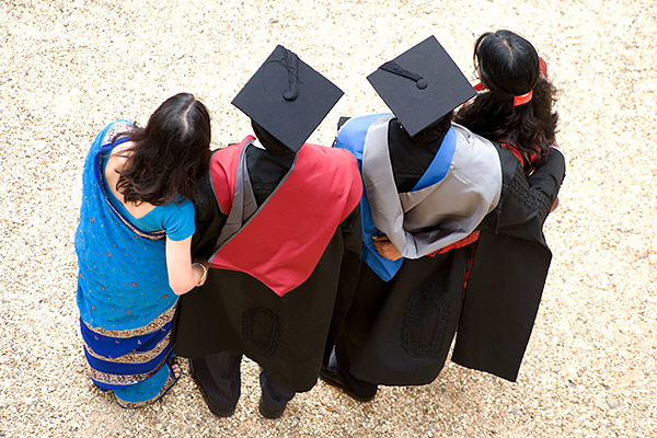 Two graduating students wearing academic dress stand outside with family members