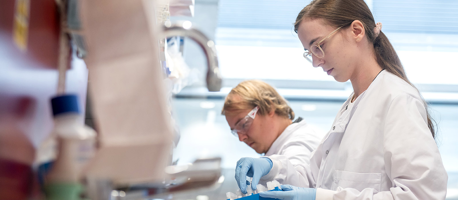 Two researchers in profile work in the lab, handling test tubes with brightly coloured red and blue tops