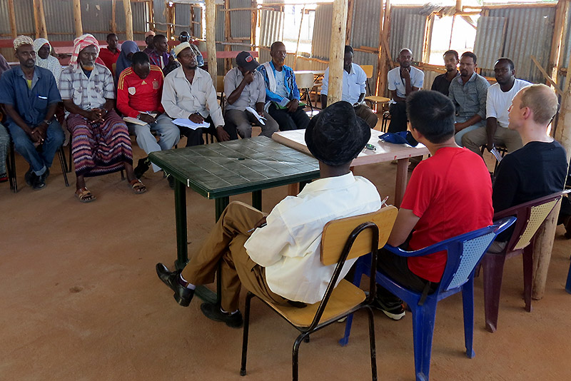 Alex Betts sits with refugees in a semi-circle on chairs to conduct a focus group 