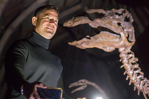 A smiling Steve Backshall explores OUMNH after dark, torch in hand, as he views a dinosaur skeleton
