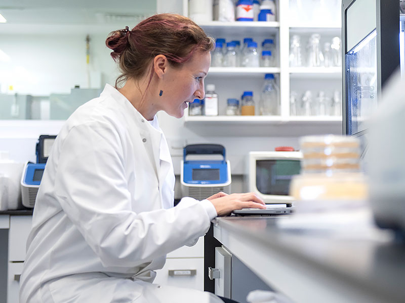 Professor Emma Slack wears a white lab coat and sits at a laptop in the lab