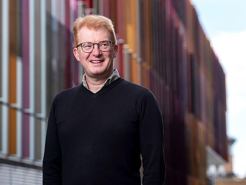 Professor Matt Higgins stands in front of the Dorothy Crowfoot Hodgkin Building, which is covered with red, pink and orange panels