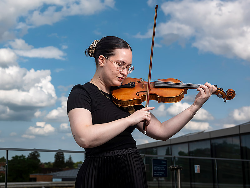 Carmen Jorge Díaz plays the violin on the roof of the Mathematical Institute against a blue sky and white clouds