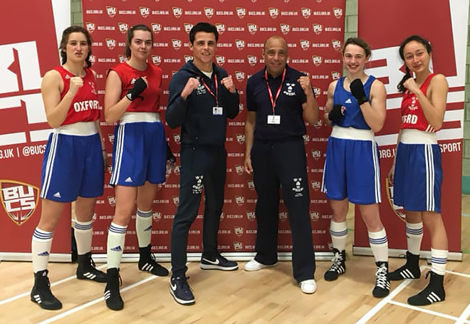 Photo of four female boxers stood along side coaches all facing the camera with their fists up and smiling