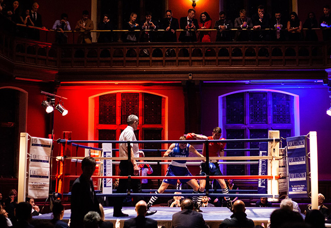 Photo of a live boxing match staged in a college hall with spectators sat around the ring and above stading on a balcony