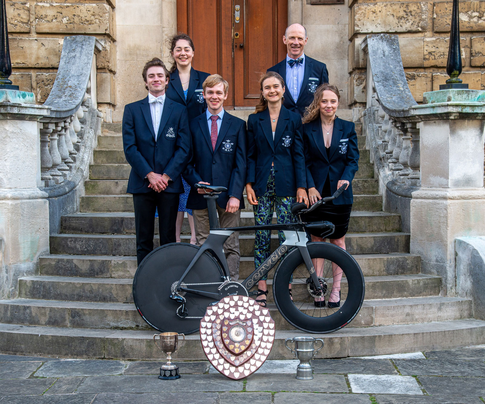 The six 2021/22 road cycling Varsity champions in club blazers on the steps to the Radcliffe Camera, posing with Varsity trophies & a time-trial bike.