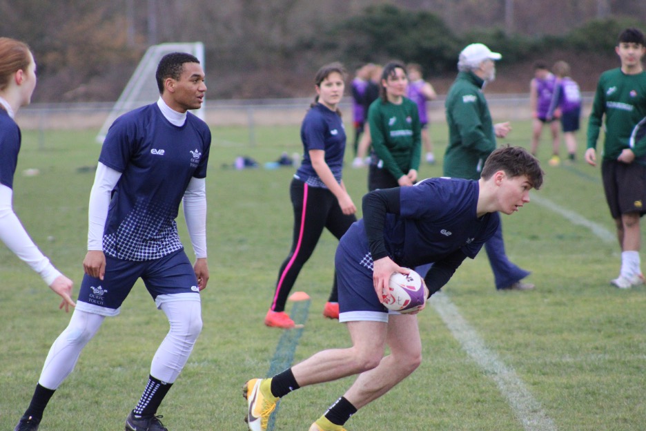 A male player in Oxford dark blue kit taps off the ball to start the game. Another male player trails in preparation for the next possession. Two female players run passing lines against the opposition.